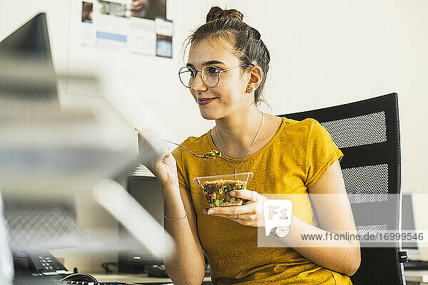 Lächelnde Geschäftsfrau  die Essen isst  während sie im Büro am Computer sitzt