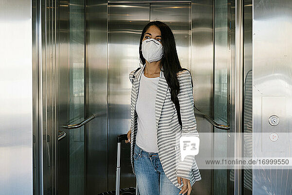 Frau mit Schutzmaske geht mit Gepäck aus dem Aufzug am Bahnhof