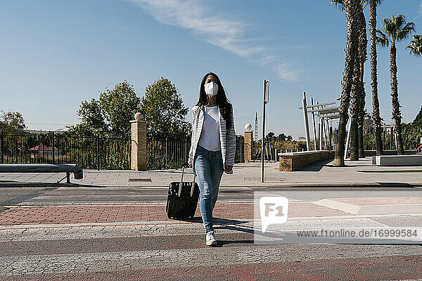 Frau mit Gepäck überquert Straße gegen blauen Himmel an einem sonnigen Tag während der Pandemie