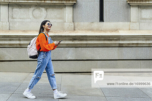 Lächelnde Frau mit Rucksack und Mobiltelefon  die beim Gehen auf dem Fußweg wegschaut