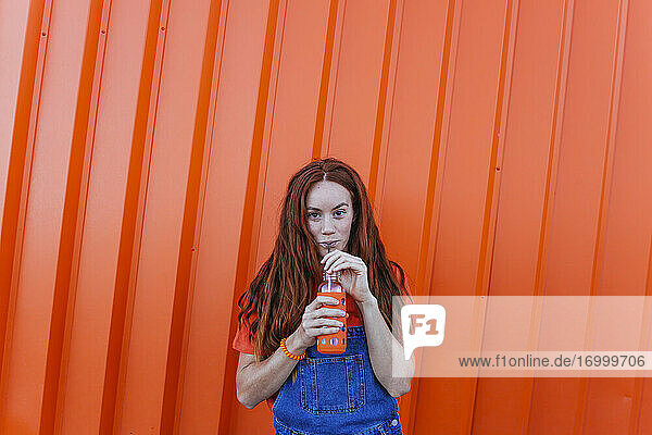 Rothaarige Frau trinkt Saft  während sie gegen eine orangefarbene Wand steht