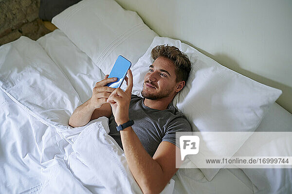 Lächelnder Mann, der ein Smartphone benutzt, während er auf dem Bett im Schlafzimmer liegt