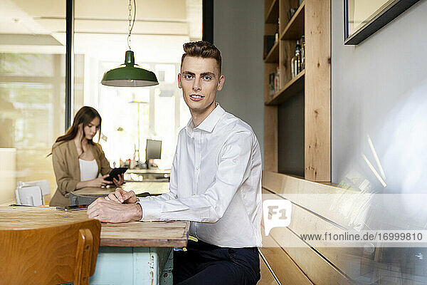 Junger Geschäftsmann sitzt auf einem Tisch mit einem Kollegen  der im Hintergrund im Büro arbeitet