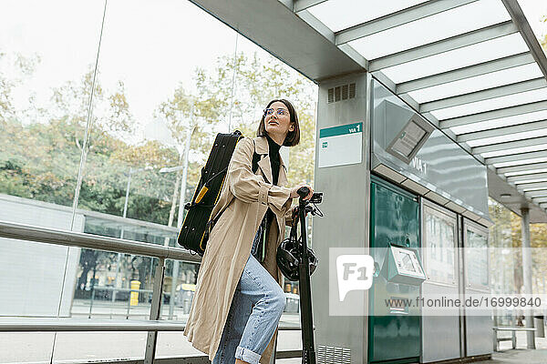 Lächelnde Frau mit Tretroller und Instrumentenkoffer an der Straßenbahn in der Stadt