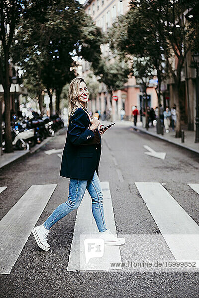 Smiling blond woman holding mobile phone crossing street in city