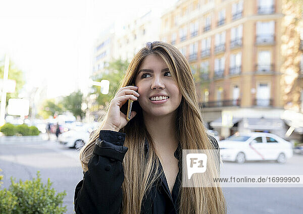 Lächelnde Frau  die auf der Straße in der Stadt steht und mit ihrem Handy telefoniert