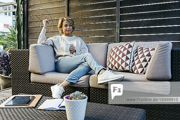 Blonde Frau schaut weg und hält eine Kaffeetasse auf dem Sofa im Dachgarten