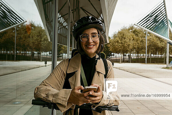 Lächelnde Frau mit Fahrradhelm  die mit einem Elektroroller auf dem Gehweg steht