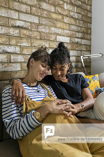 Lesbian couple sitting with hands clasped on stomach while sitting at home