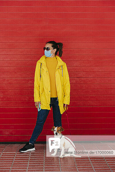 Frau mit Gesichtsmaske und Hund  in gelbem Regenmantel vor roter Wand
