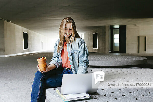 Lächelnde blonde Studentin  die auf einen Laptop schaut  während sie mit einem wiederverwendbaren Kaffeebecher auf dem Campus sitzt