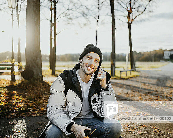 Lächelnder Mann  der sein Smartphone hält  während er im Herbst im Park sitzt