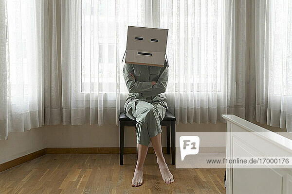Frau  die einen Karton auf dem Kopf mit einem gelangweilten Smiley trägt und mit verschränkten Armen auf einem Stuhl sitzt