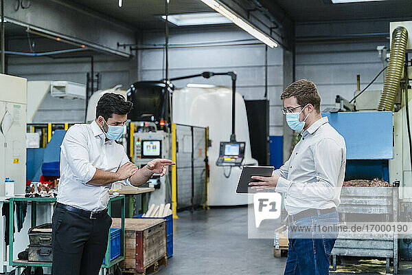 Male coworkers using digital tablet while maintaining social distancing in factory
