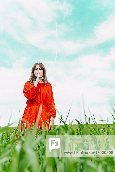 Portrait of woman wearing red dress standing in a field with finger on mouth