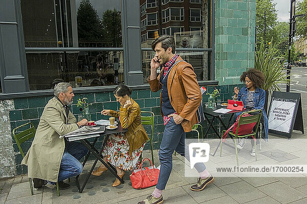 Geschäftsmann  der mit seinem Smartphone telefoniert  während er an Kunden vorbeigeht  die in einem Straßencafé sitzen