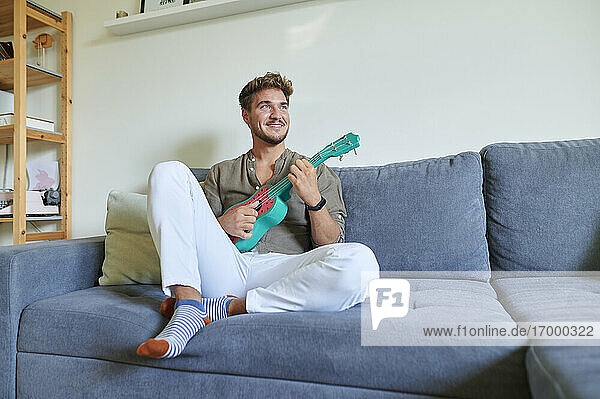 Lächelnder männlicher Musiker spielt Ukulele  während er zu Hause auf dem Sofa sitzt