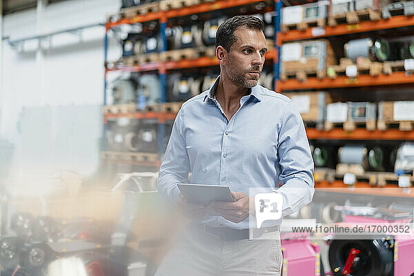 Male entrepreneur holding digital tablet looking away while standing at factory