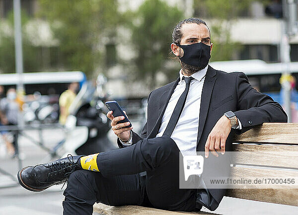 Porträt eines Geschäftsmannes mit Gesichtsschutzmaske  der auf einer Bank sitzt und ein Smartphone in der Hand hält