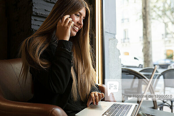 Lächelnde Frau mit Laptop  die in einem Café sitzt und mit ihrem Handy telefoniert