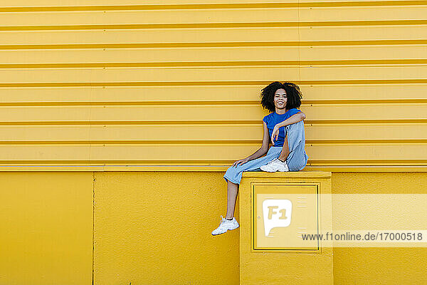 Hübsche Frau sitzt auf einem Podest vor einer gelben Wand und lächelt