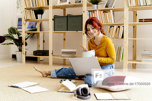 Lächelnde Frau  die einen Laptop benutzt  während sie zu Hause sitzt und isst
