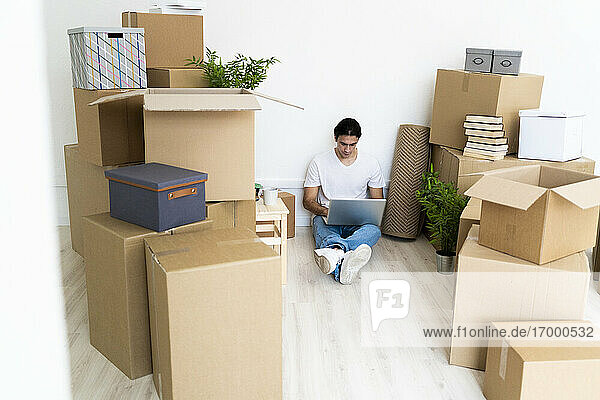 Mann arbeitet an einem Laptop  umgeben von Kisten  während er in seiner neuen Wohnung sitzt