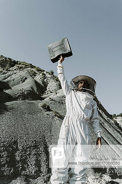 Mann in Imkerkleid hält Aktentasche in einer trockenen apokalyptischen Landschaft hoch