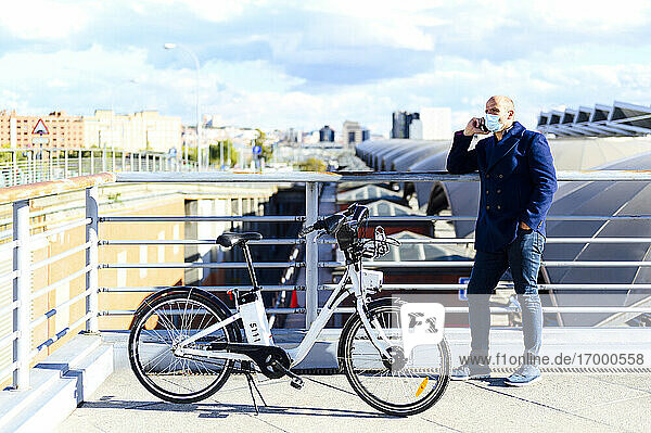 Mann spricht in sein Smartphone  während er mit der Hand in der Tasche auf einer Brücke während COVID-19 steht