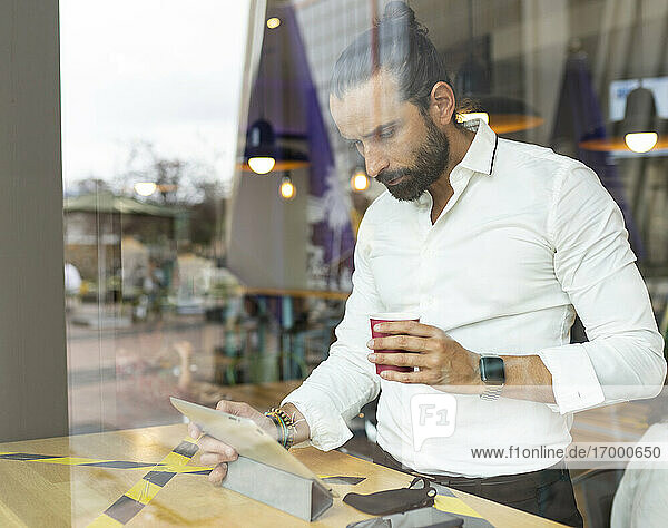 Unverfälschtes Porträt eines bärtigen Geschäftsmannes  der vor einem digitalen Tablet in einem Café Kaffee trinkt