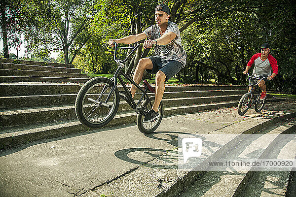 Junger Mann macht Wheelie-Stunt mit BMX-Fahrrad gegen einen Freund  der auf den Stufen eines öffentlichen Parks radelt