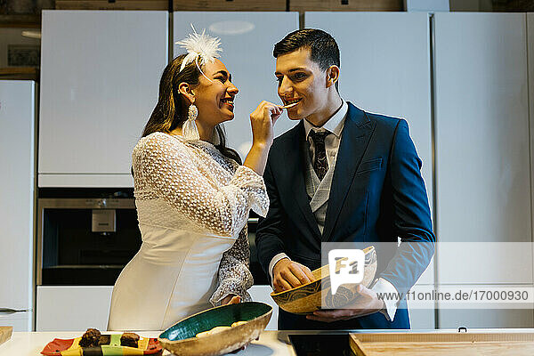 Lächelnde Braut  die den Bräutigam mit Kartoffelchips füttert  während sie in der Küche zu Hause steht
