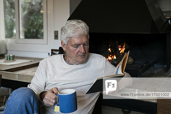 Älterer Mann mit Kaffeetasse  der ein Buch liest  während er zu Hause am Kamin sitzt