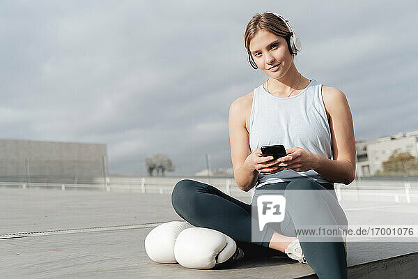 Sportswoman wearing headphones using mobile phone while sitting outdoors