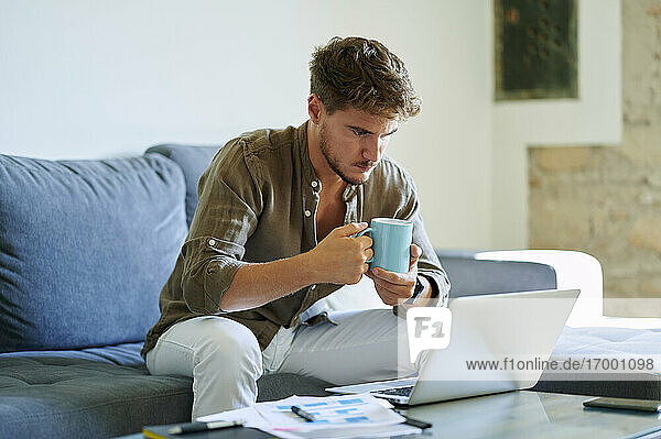 Junger männlicher Unternehmer hält Kaffeetasse  während er im Wohnzimmer am Laptop arbeitet