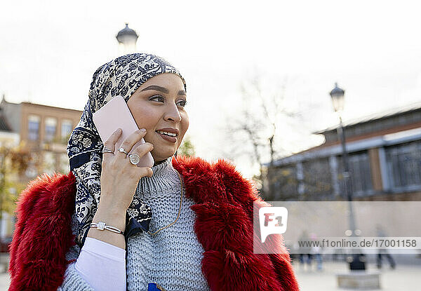 Beautiful woman wearing headscarf and fur coat talking on mobile phone while standing in city