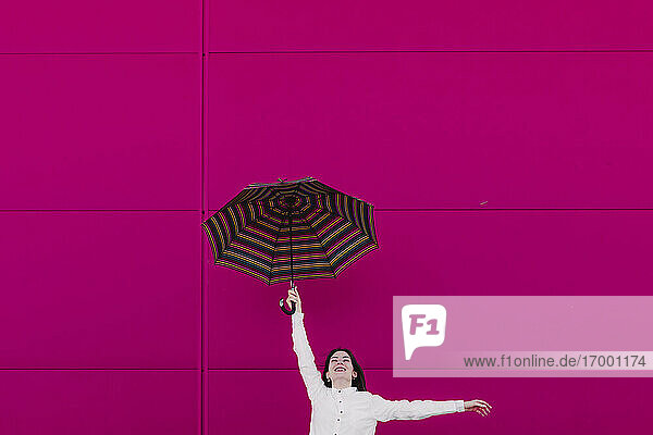 Junge Frau springt mit Regenschirm vor einer rosa Wand