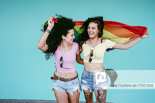 Cheerful lesbian girlfriends holding rainbow flag while standing against wall