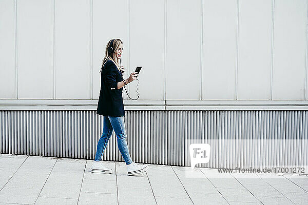 Woman listening music through headphones while walking on sidewalk in city