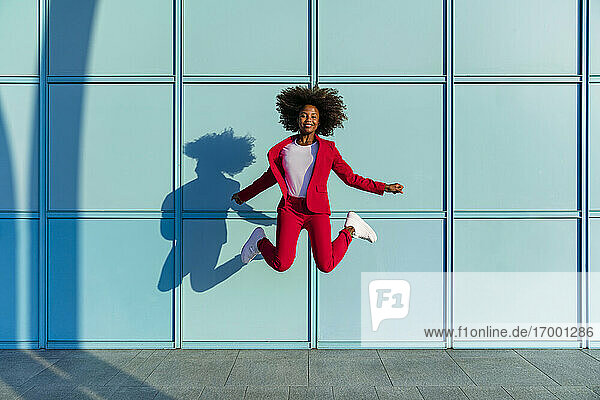 Lächelnde Frau springt gegen blaue Wand an einem sonnigen Tag