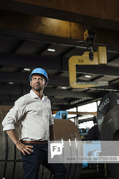 Confident businessman with hands on hip looking up while standing in factory