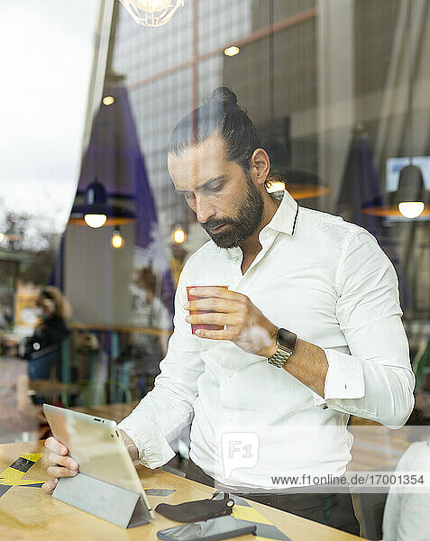 Unverfälschtes Porträt eines bärtigen Geschäftsmannes  der vor einem digitalen Tablet in einem Café Kaffee trinkt