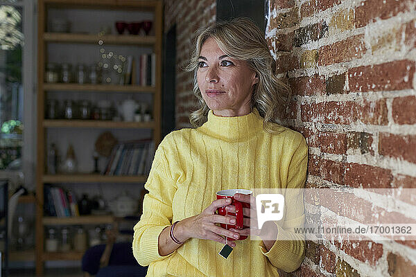 Reife Frau mit Teetasse  die wegschaut  während sie sich an eine Backsteinmauer in der Küche lehnt