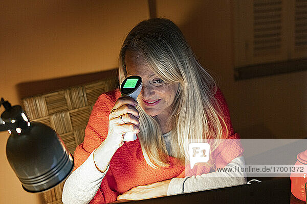Lächelnde ältere Frau  die ein Infrarot-Thermometer benutzt  während sie einen Videoanruf auf einem Laptop zu Hause betrachtet