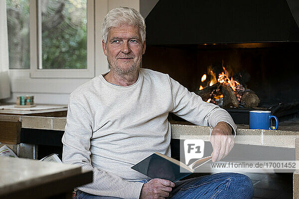 Lächelnder älterer Mann  der ein Buch liest  während er zu Hause am Kamin sitzt