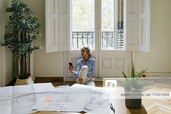 Geschäftsfrau  die ein Smartphone benutzt  während sie auf dem Boden vor dem Fenster im Büro sitzt