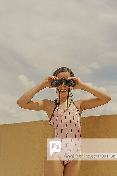 Portrait of laughing girl in swimsuit standing on roof terrace looking through binoculars