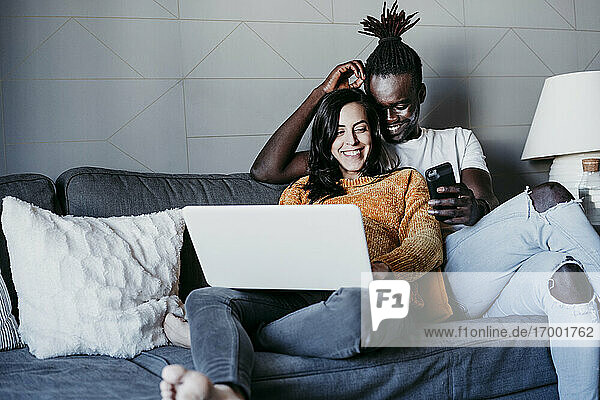 Glücklicher junger Mann mit schwangerer Frau  die auf ihr Mobiltelefon schaut  während sie zu Hause einen Laptop benutzen