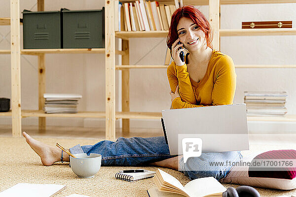 Lächelnde Frau  die mit dem Handy telefoniert  während sie zu Hause am Laptop sitzt