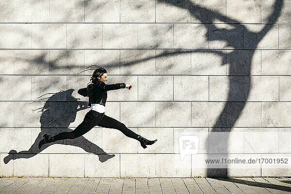 Junge Frau macht einen großen Sprung auf einer Mauer mit einem Schatten eines Baumes